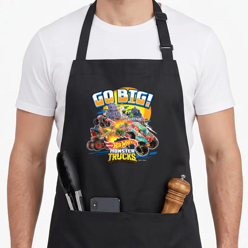 Hobby truck aprons - GO BIG! text over monster trucks, palm trees, and Hot Wheels logo in yellow blue red green — shown on a black cotton canvas apron against a white background