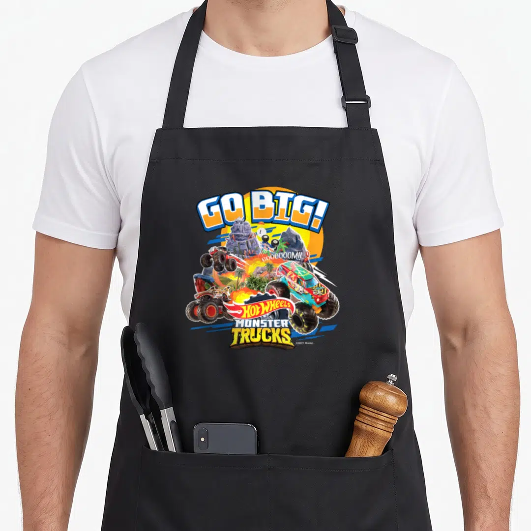 Hobby truck aprons - GO BIG! text over monster trucks, palm trees, and Hot Wheels logo in yellow blue red green — shown on a black cotton canvas apron against a white background
