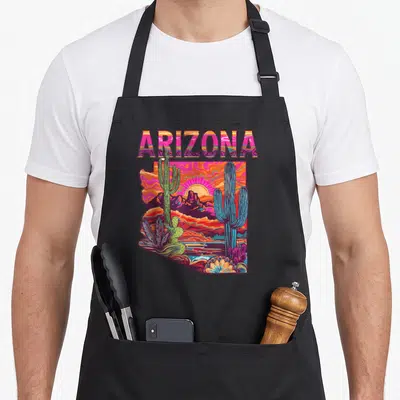 Flag desert cactus apron - Retro ARIZONA text above a state-shaped desert scene with cacti in pink orange purple blue — shown on a black cotton canvas apron against a white background