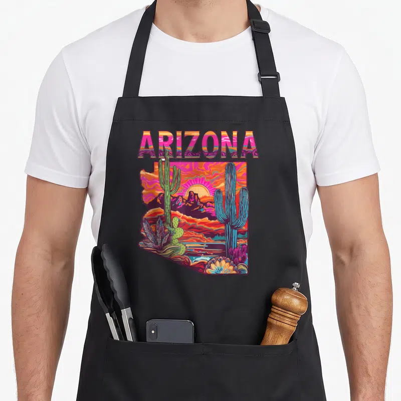 Flag desert cactus apron - Retro ARIZONA text above a state-shaped desert scene with cacti in pink orange purple blue — shown on a black cotton canvas apron against a white background