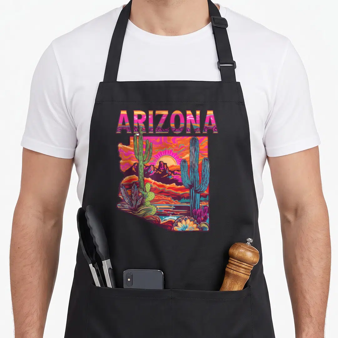 Flag desert cactus apron - Retro ARIZONA text above a state-shaped desert scene with cacti in pink orange purple blue — shown on a black cotton canvas apron against a white background