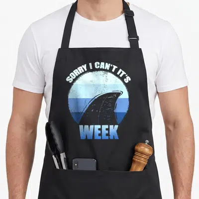 Sorry shark apron - Grey shark fin in blue gradient circle with white text SORRY I CAN'T IT'S WEEK — shown on a black cotton canvas apron against a white background