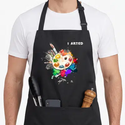 Artist colorful aprons - I ARTED text above watercolor palette and multicolored paint splatters with brushes — shown on a black cotton canvas apron against a white background