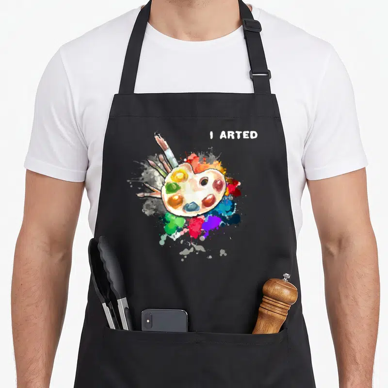Artist colorful aprons - I ARTED text above watercolor palette and multicolored paint splatters with brushes — shown on a black cotton canvas apron against a white background