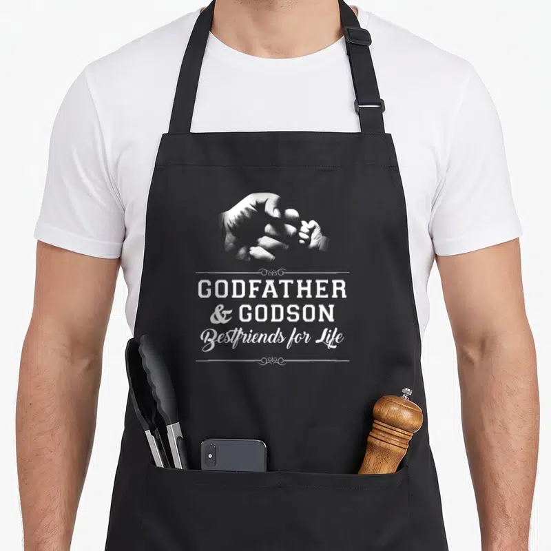 Bump friends apron - White hands fist bumping above stacked text reading GODFATHER and GODSON Bestfriends for Life — shown on a black cotton canvas apron against a white background