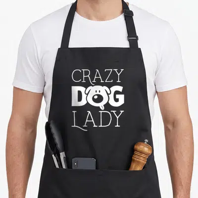 Pup cooking apron - White CRAZY DOG LADY text with dog head replacing the O on black fabric — shown on a black cotton canvas apron against a white background