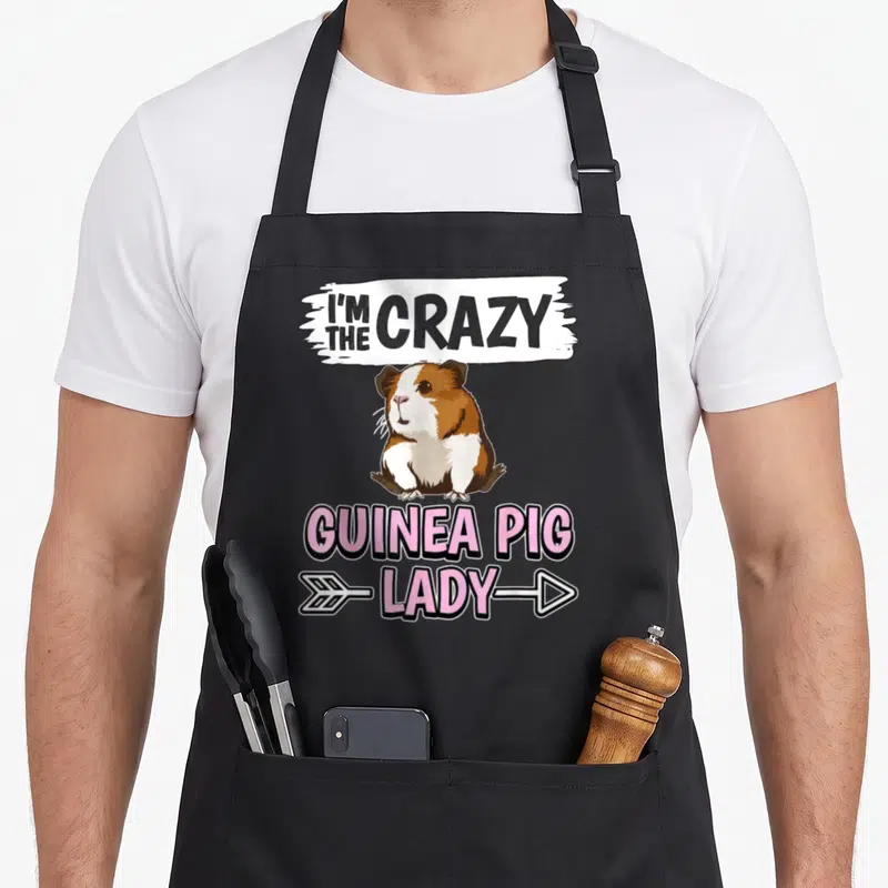 Cavy pig crazy guinea lady apron - Cartoon guinea pig in brown and white with pink and grey text reading I'm the crazy — shown on a black cotton canvas apron against a white background