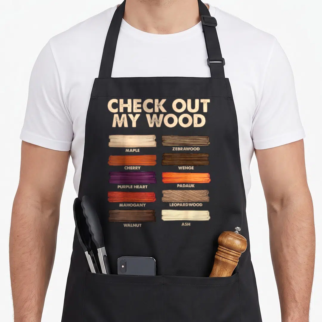 Woodworking carpentry apron - Check Out My Wood text above ten labeled wood samples in beige brown orange and purple — shown on a black cotton canvas apron against a white background