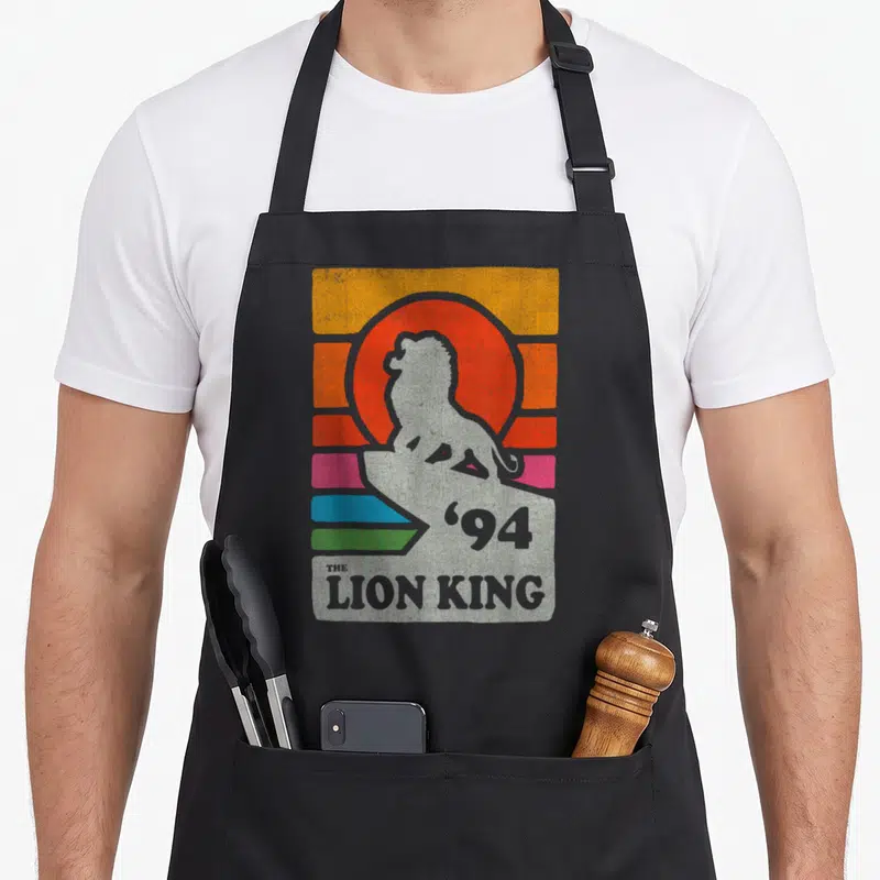 Pride line apron - Grey lion on rock with '94 THE LION KING' text over orange sun and colored stripes — shown on a black cotton canvas apron against a white background