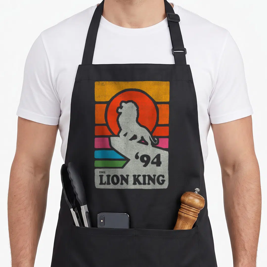 Pride line apron - Grey lion on rock with '94 THE LION KING' text over orange sun and colored stripes — shown on a black cotton canvas apron against a white background