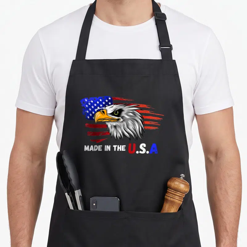 Bald tattoo apron - Cartoon eagle head over brush stroke flag with Made in the U.S.A text — shown on a black cotton canvas apron against a white background
