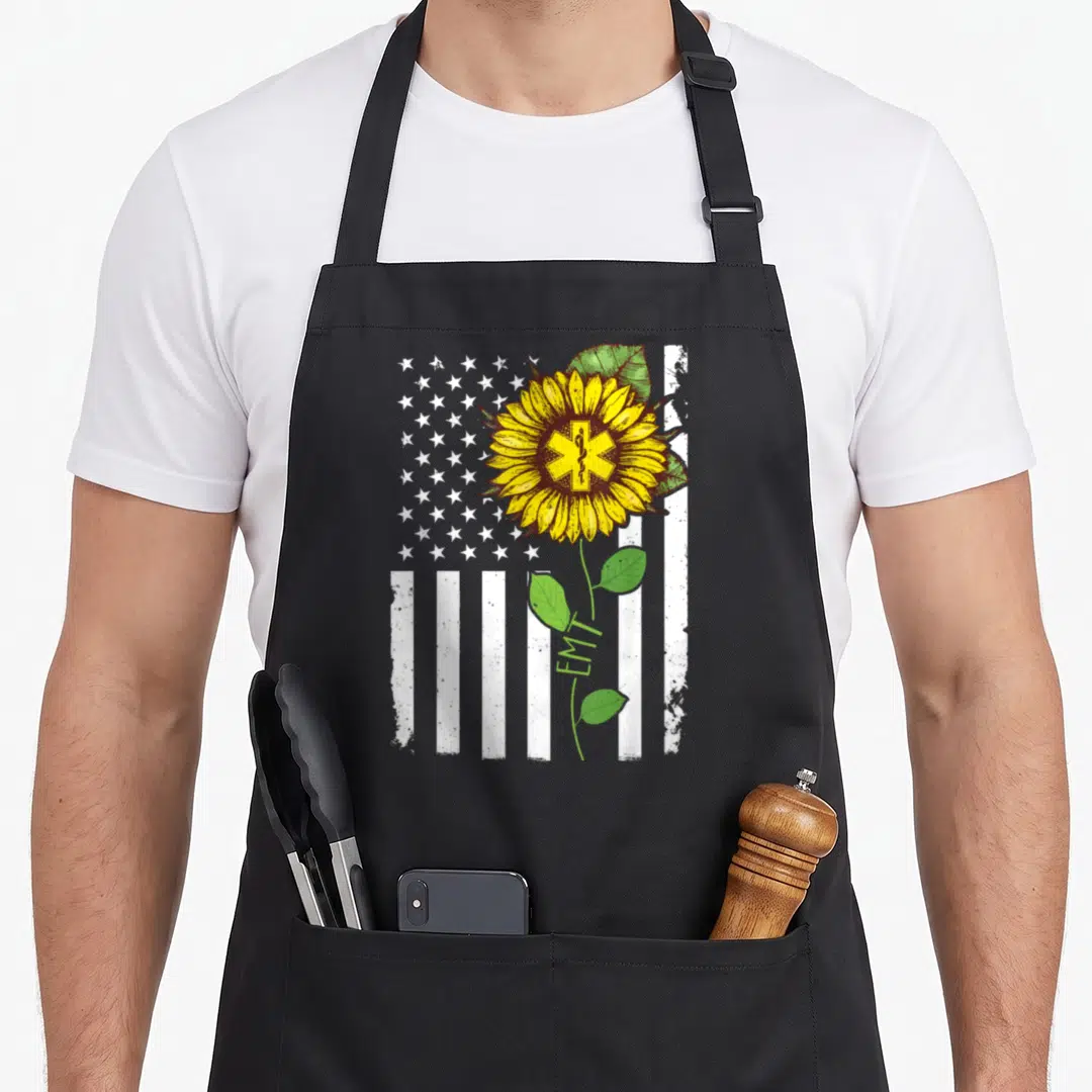Sunflower american flag apron - Yellow sunflower with Star of Life over distressed white flag, vertical EMT text — shown on a black cotton canvas apron against a white background