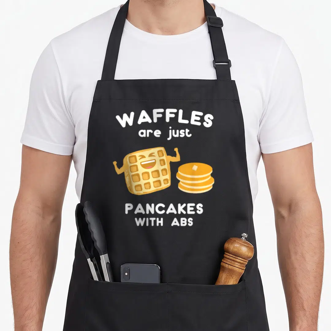Pun pancake apron - Cartoon waffle flexing muscles next to pancakes with text WAFFLES are just PANCAKES WITH ABS — shown on a black cotton canvas apron against a white background