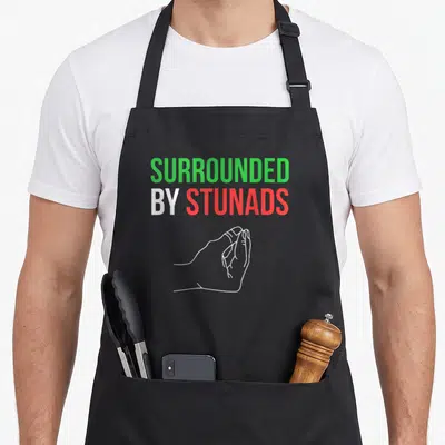 Surrounded italian apron - Text SURROUNDED BY STUNADS with green white red hand gesture line art — shown on a black cotton canvas apron against a white background