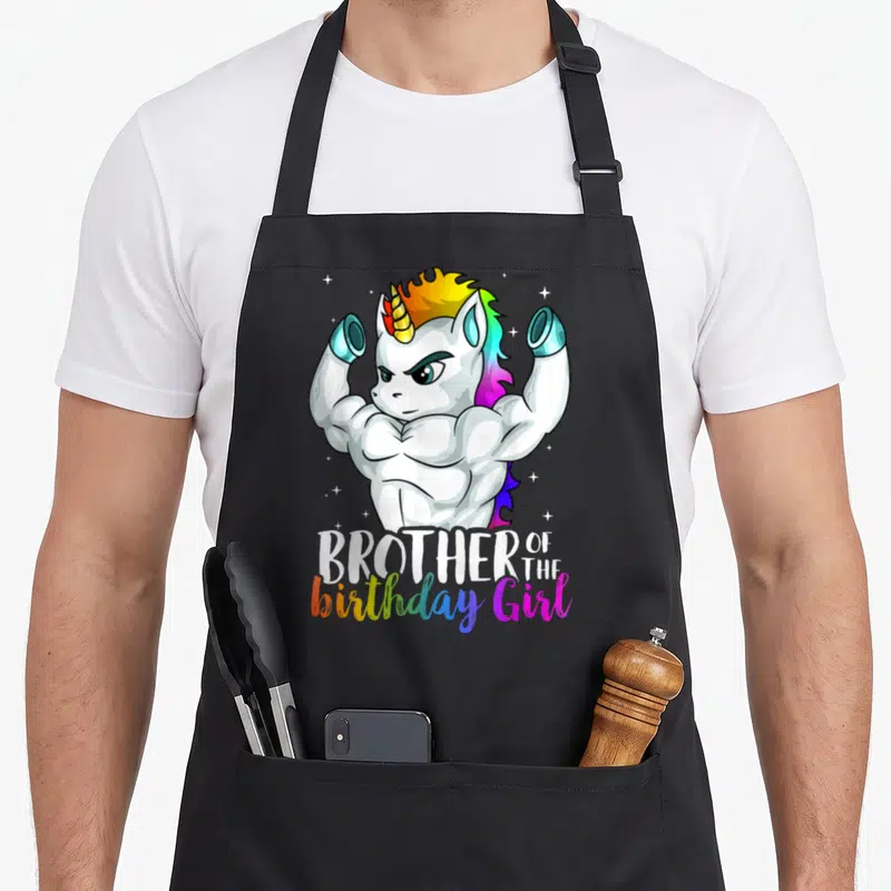 Grandkid birthday apron - Muscular white unicorn flexing arms above rainbow text reading Brother of the birthday Girl — shown on a black cotton canvas apron against a white background