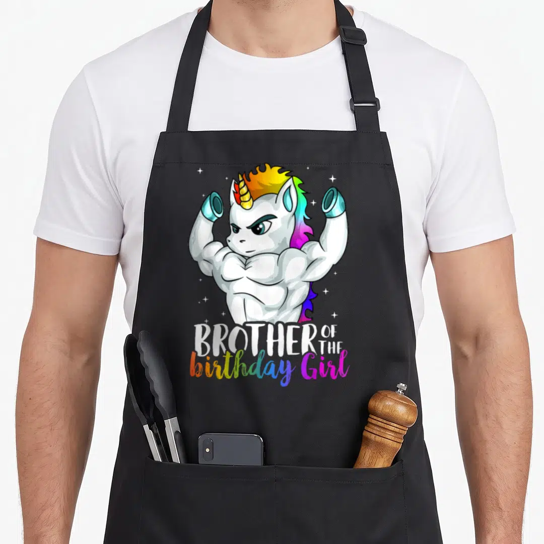 Grandkid birthday apron - Muscular white unicorn flexing arms above rainbow text reading Brother of the birthday Girl — shown on a black cotton canvas apron against a white background