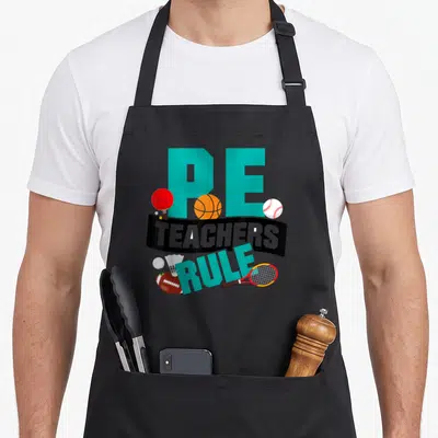 Rule teacher apron - PE TEACHERS RULE text with basketball, racket, football, and baseball in cartoon style — shown on a black cotton canvas apron against a white background