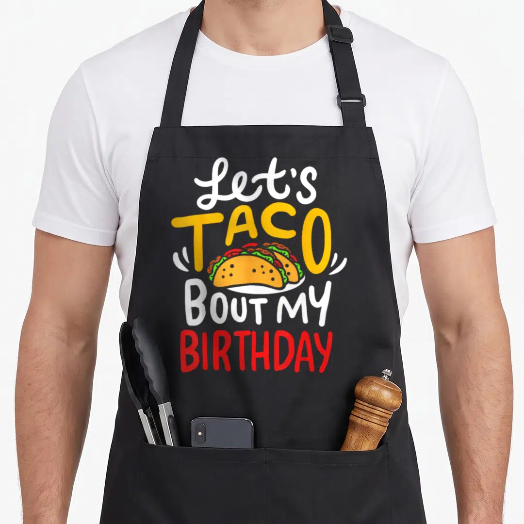 Taco birthday apron - Let's Taco Bout My Birthday text with cartoon tacos in white yellow red and green — shown on a black cotton canvas apron against a white background