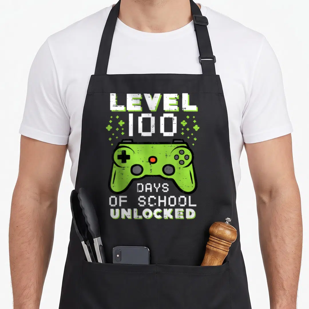 Gamer day level apron - Pixel art green game controller with white text Level 100 Days Of School Unlocked and small — shown on a black cotton canvas apron against a white background