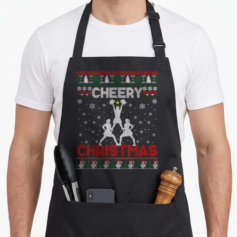 Cheering ugly sweater apron - Retro knit cheerleader pyramid with CHEERY CHRISTMAS text and snowflake borders — shown on a black cotton canvas apron against a white background