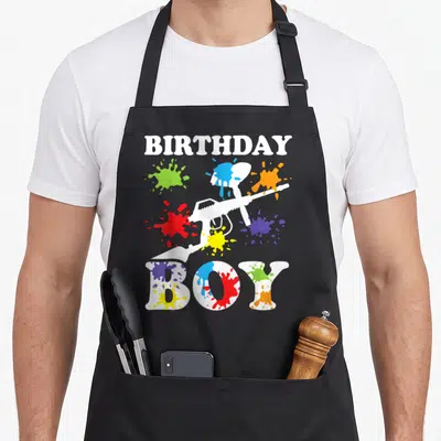 Cute paintball birthday apron - BIRTHDAY BOY text with white toy gun and colorful paint splatters on black bib — shown on a black cotton canvas apron against a white background