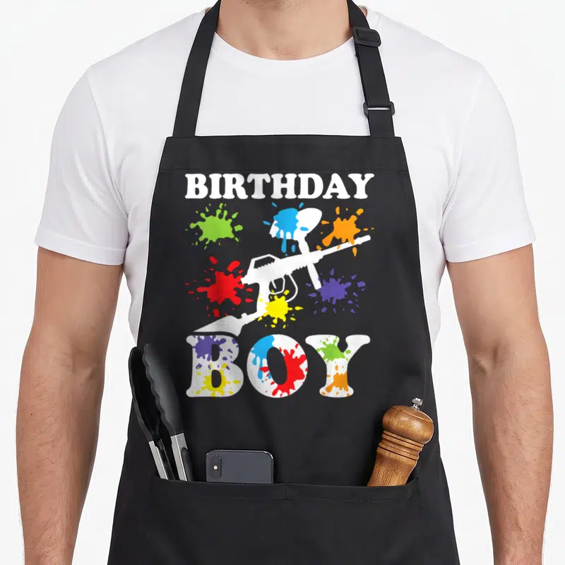 Cute paintball birthday apron - BIRTHDAY BOY text with white toy gun and colorful paint splatters on black bib — shown on a black cotton canvas apron against a white background