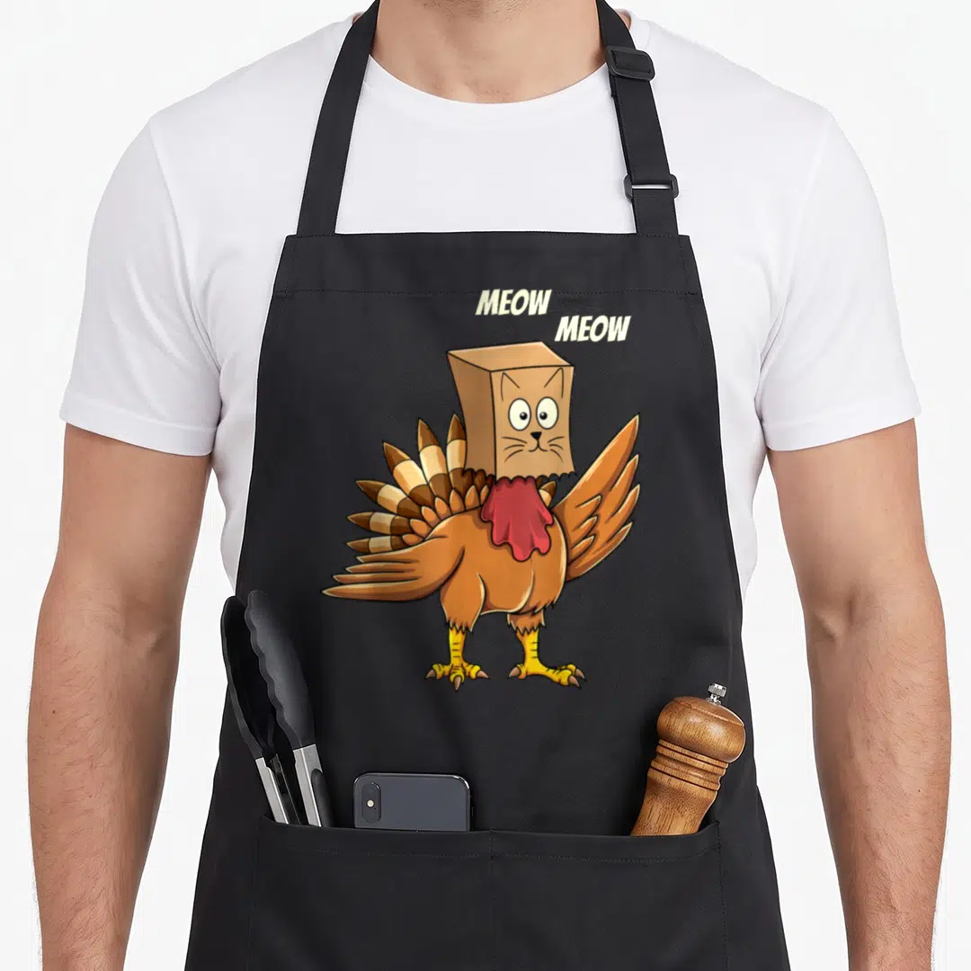 Meow turkey apron - Cartoon turkey wearing a cardboard cat mask with MEOW MEOW text floating above — shown on a black cotton canvas apron against a white background