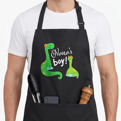 Nanas dinosaur apron - Two green cartoon dinosaurs flanking white script reading Nana's boy! — shown on a black cotton canvas apron against a white background
