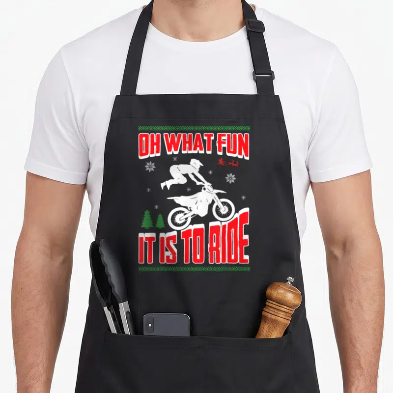 Dirt xmas apron - Motocross rider jumping between OH WHAT FUN IT IS TO RIDE text with snowflakes and trees — shown on a black cotton canvas apron against a white background