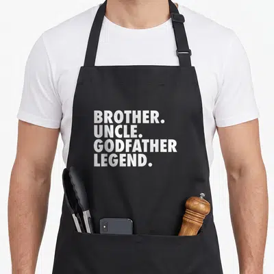 Godchild goddaughter apron - White stacked text reading BROTHER UNCLE GODFATHER LEGEND with periods — shown on a black cotton canvas apron against a white background