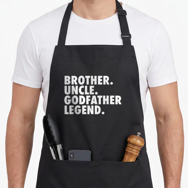 Godchild goddaughter apron - White stacked text reading BROTHER UNCLE GODFATHER LEGEND with periods — shown on a black cotton canvas apron against a white background