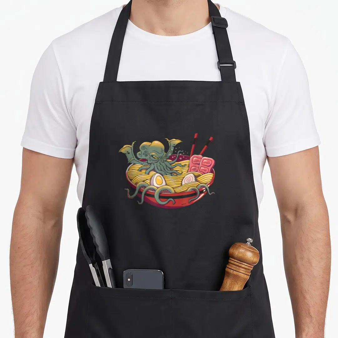Ramen cthulhu apron - Green octopus in red ramen bowl with yellow noodles, pink fish cake, and chopsticks — shown on a black cotton canvas apron against a white background