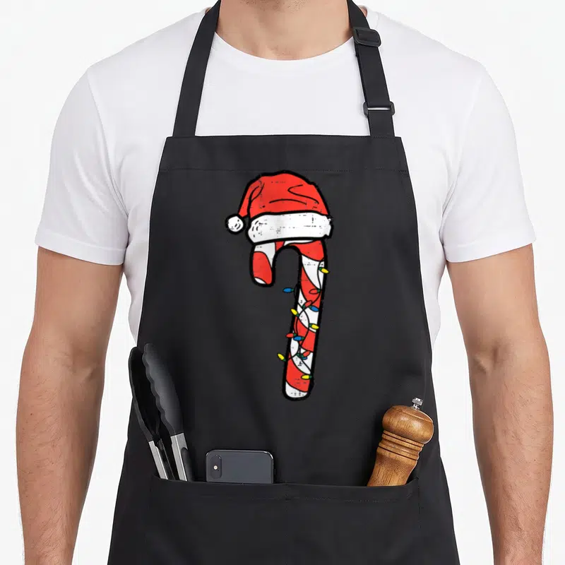 Santa candy cane apron - Cartoon candy cane with Santa hat and colorful Christmas lights on black fabric — shown on a black cotton canvas apron against a white background