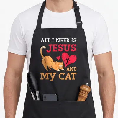 Jesus kitty cat apron - Orange cat stretching beside red heart with black cross under stacked white and red text — shown on a black cotton canvas apron against a white background