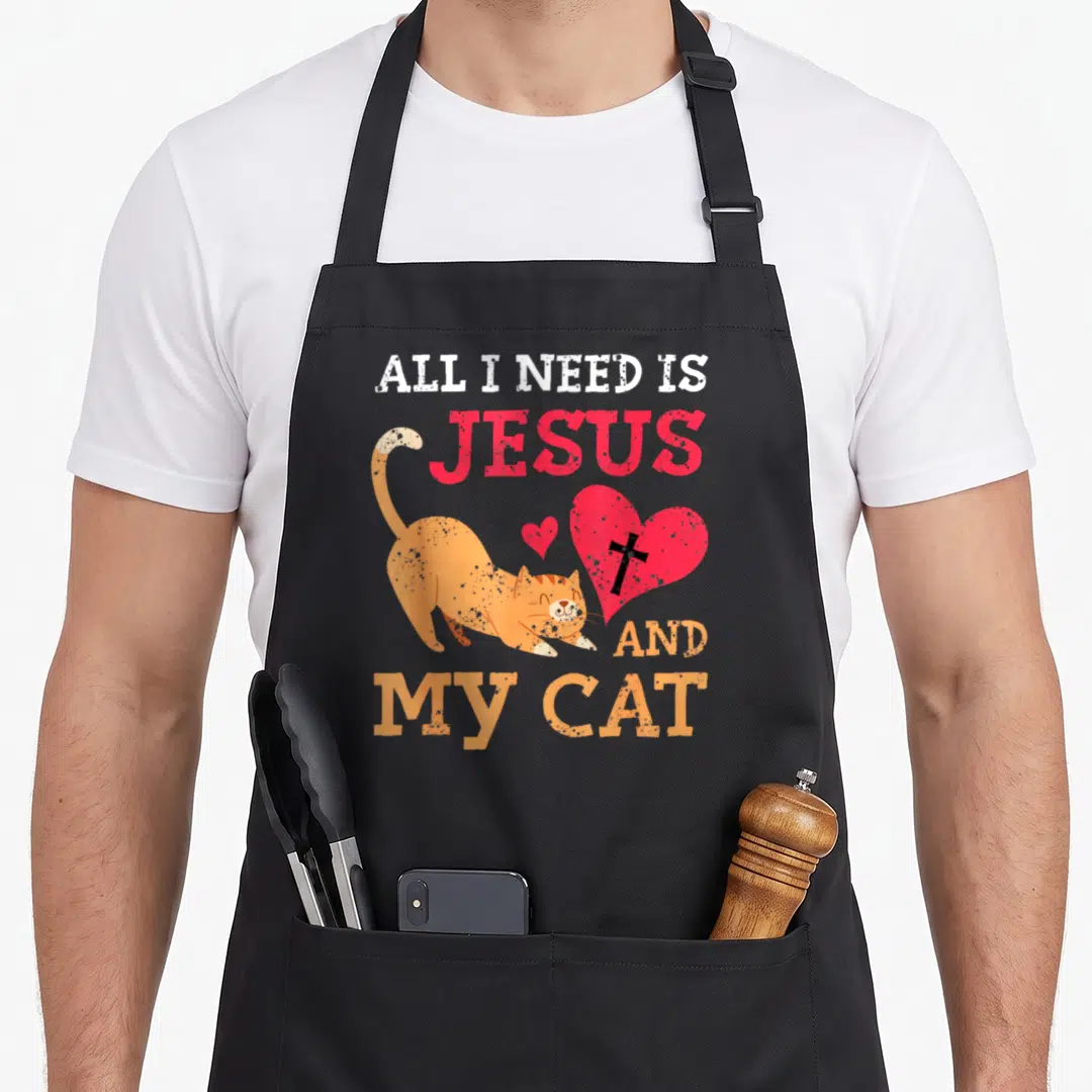 Jesus kitty cat apron - Orange cat stretching beside red heart with black cross under stacked white and red text — shown on a black cotton canvas apron against a white background