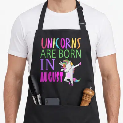 Dab birthday apron - Stacked text reading UNICORNS ARE BORN IN AUGUST beside a dabbing rainbow-maned unicorn — shown on a black cotton canvas apron against a white background