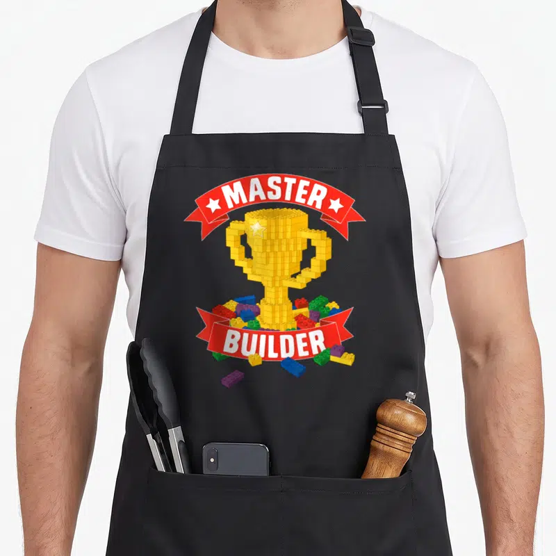 Building block grill master apron - Yellow block trophy with red ribbons reading MASTER BUILDER surrounded by scattered — shown on a black cotton canvas apron against a white background