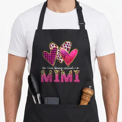 Called valentine's day apron - Three patterned hearts above love being called MIMI text in pink gold black white — shown on a black cotton canvas apron against a white background
