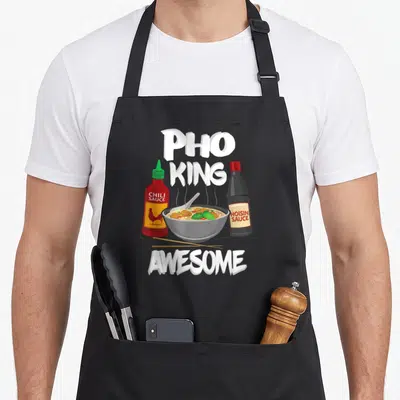 King pho apron - Cartoon pho bowl with chili sauce, hoisin sauce, and chopsticks lying in front under stacked white text — shown on a black cotton canvas apron against a white background