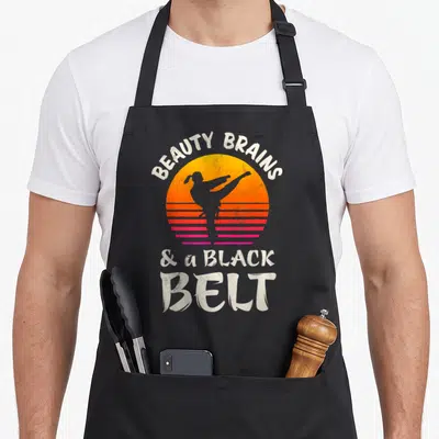 Brain beauty apron - Retro sunset circle with kicking martial artist silhouette and white arched text — shown on a black cotton canvas apron against a white background