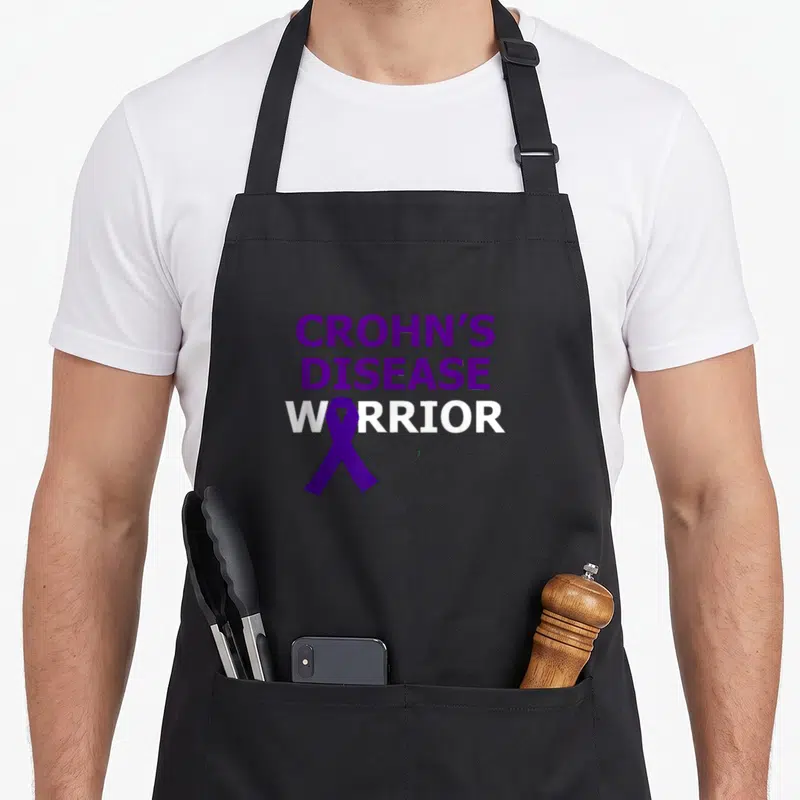 Crohn faith aprons - Purple and white text CROHN'S DISEASE WARRIOR with ribbon replacing the O letter — shown on a black cotton canvas apron against a white background
