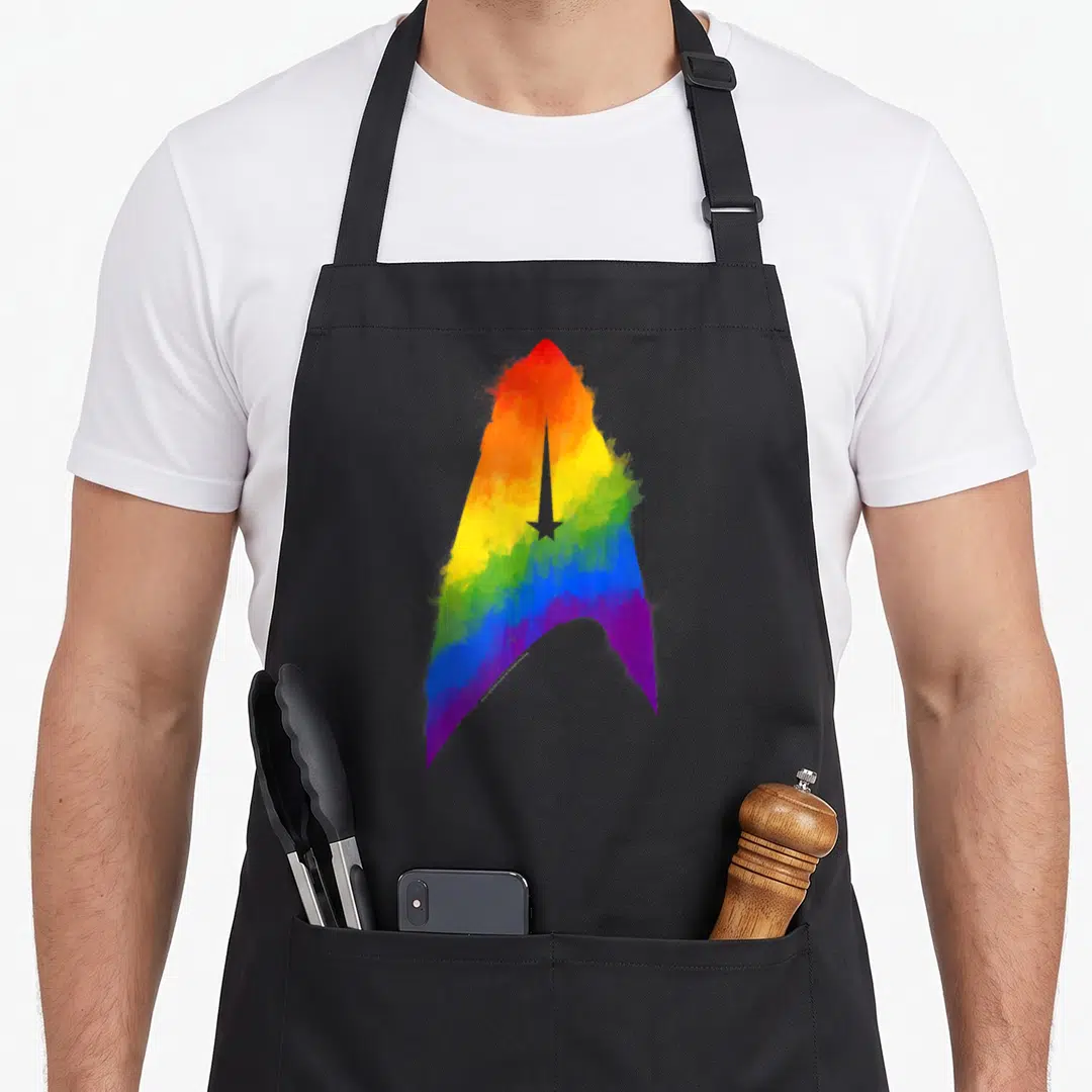 Discovery rainbow apron - Watercolor rainbow gradient Starfleet delta insignia with central star cutout — shown on a black cotton canvas apron against a white background