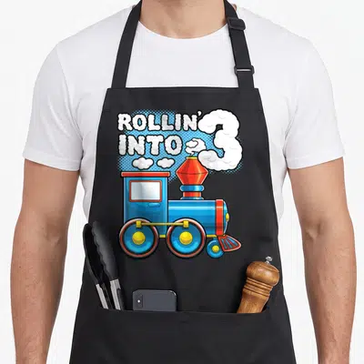 Rollin train birthday apron - Blue steam train with smoke forming number 3 and bubble text ROLLIN INTO 3 — shown on a black cotton canvas apron against a white background