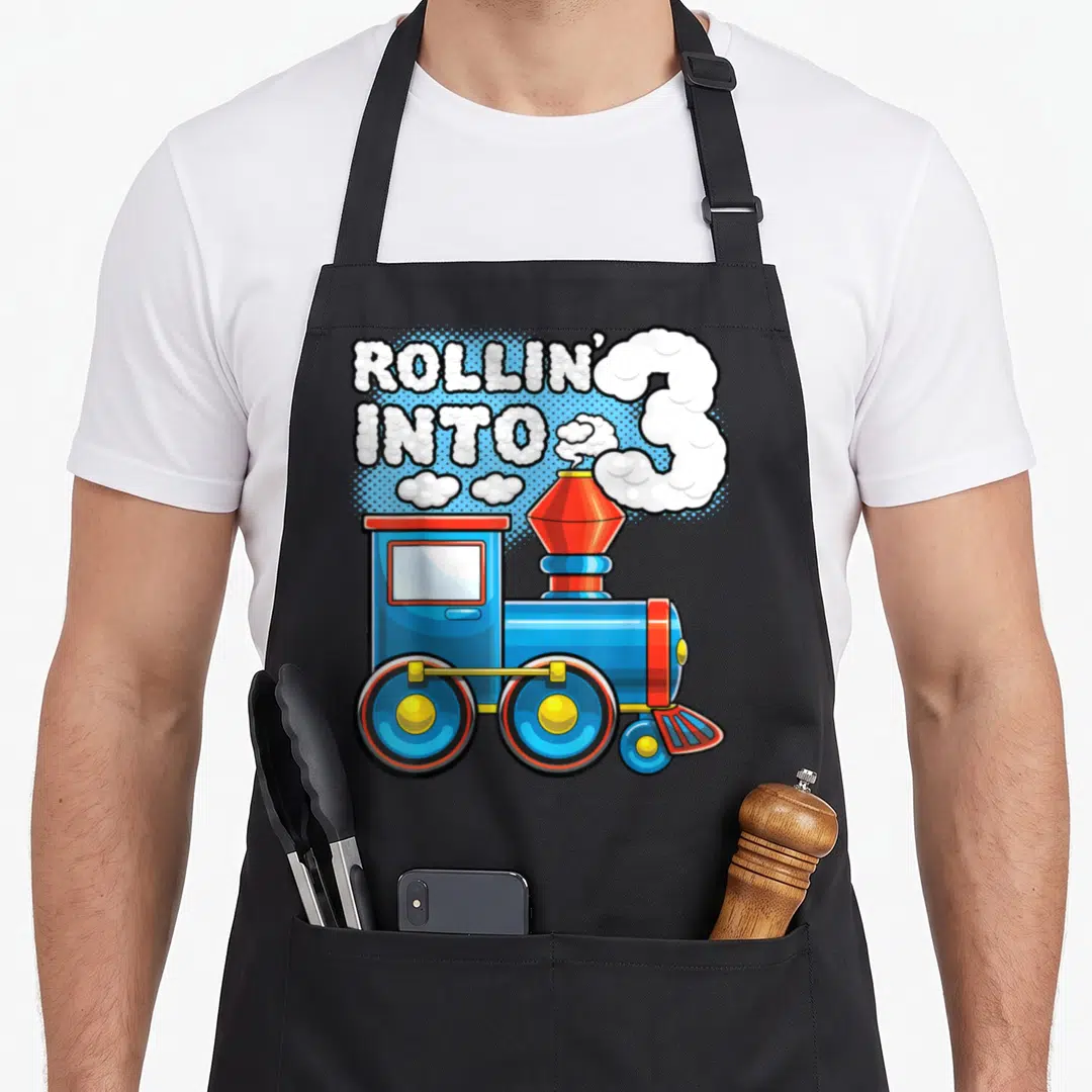 Rollin train birthday apron - Blue steam train with smoke forming number 3 and bubble text ROLLIN INTO 3 — shown on a black cotton canvas apron against a white background