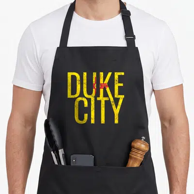 Albuquerque duke mexico apron - Stacked yellow DUKE CITY text with red Zia flag pattern inside letters and distressed — shown on a black cotton canvas apron against a white background