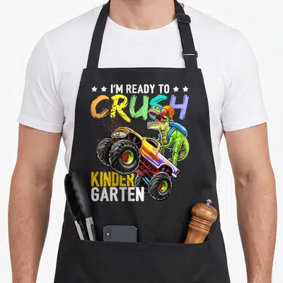 Kindergarten truck apron - T-Rex in orange cap and blue backpack driving yellow monster truck with crush kinder garten — shown on a black cotton canvas apron against a white background
