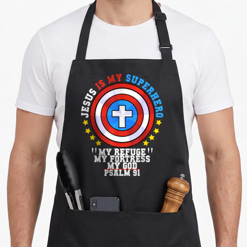 Jesus superhero apron - Circular shield with cross and yellow stars surrounded by Jesus Is My Superhero text — shown on a black cotton canvas apron against a white background
