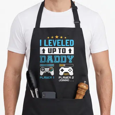 Leveled daddy apron - Stacked I Leveled Up To Daddy text with two game controllers and Player 2 Joining label — shown on a black cotton canvas apron against a white background