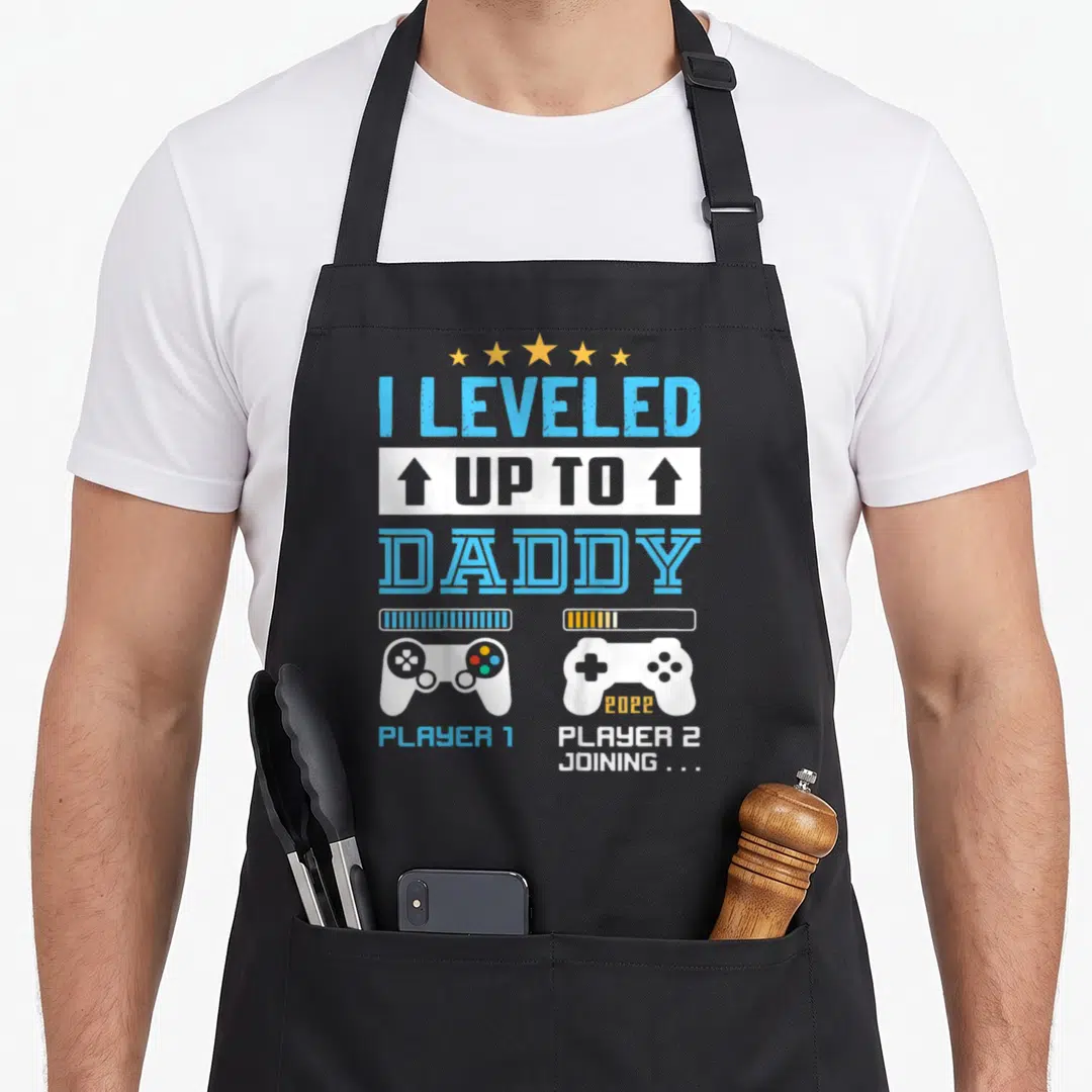 Leveled daddy apron - Stacked I Leveled Up To Daddy text with two game controllers and Player 2 Joining label — shown on a black cotton canvas apron against a white background