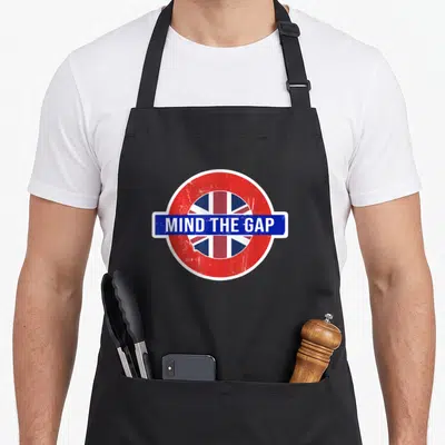 Vacation travel aprons - Retro London Underground sign with MIND THE GAP text and Union Jack flag in red white blue — shown on a black cotton canvas apron against a white background